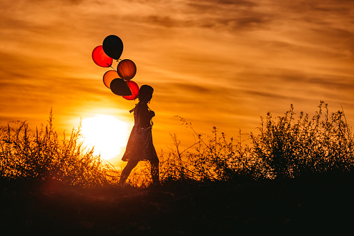 Silhouette of young woman with balloons standing on meadow at sunset.