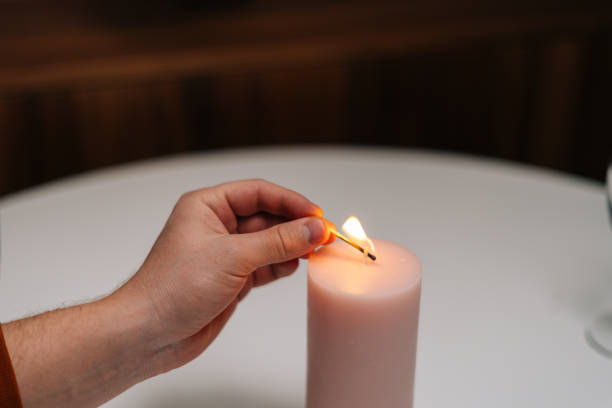 Close-up high-angle view of unrecognizable man lighting large white candle with match at table in dark living room, selective focus.