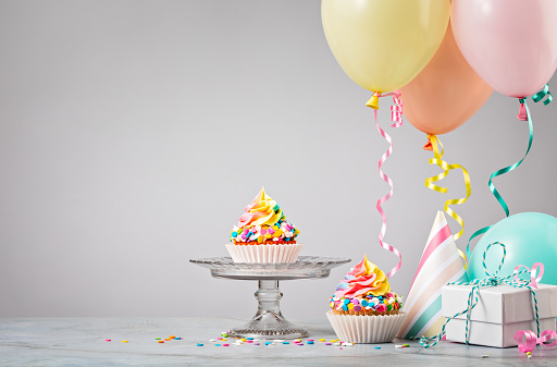 Two Rainbow Birthday cupcakes with presents, hats and colorful balloons over light grey background. Scene from a birthday party.
