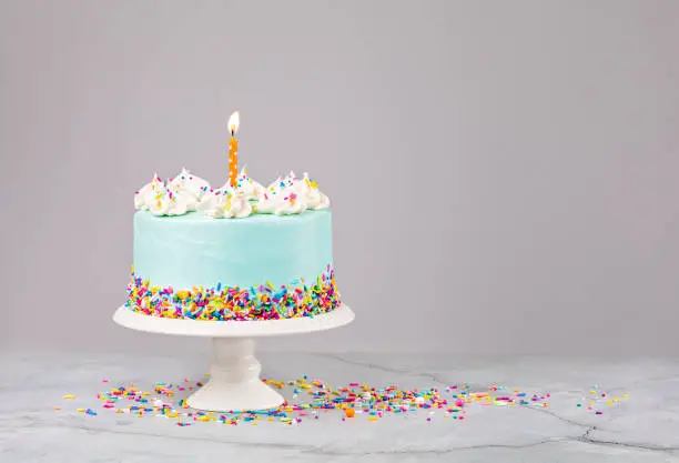 Blue Birthday Cake with colorful sprinkles and lit orange birthday candle over a light grey background.