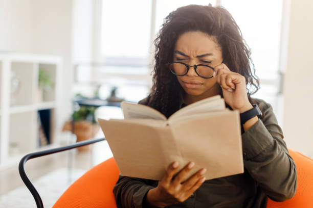 donna nera concentrata negli occhiali che cerca di leggere il libro - strizzare gli occhi foto e immagini stock