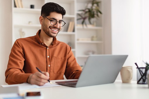 Hombre árabe sonriente con gafas usando una computadora portátil y escribiendo photo