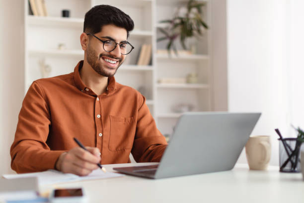 Smiling Arab man in glasses using laptop and writing stock photo