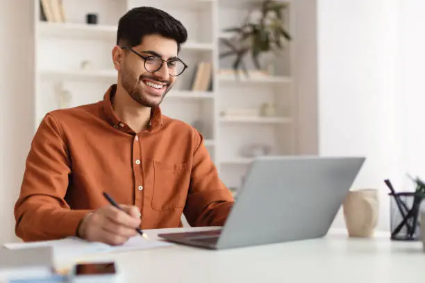 Portrait of young smiling Arab man in glasses using laptop sitting at desk, writing in notebook. Cheerful guy browsing internet, watching webinar studying online, looking at pc screen, free copy space