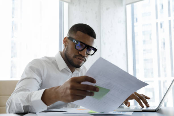 серьезный афроамериканский менеджер в стильных очках читает корпоративный документ - decisions with business men стоковые фото и изображения