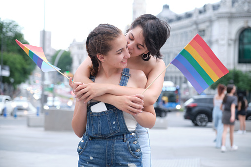 Lesbian couple hugging, smiling and kissing on the cheek to each other with lgbt flags. LGBT people.