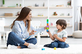 Cute little boy playing with professional teacher, sitting together on floor at nursery and building colorful tower