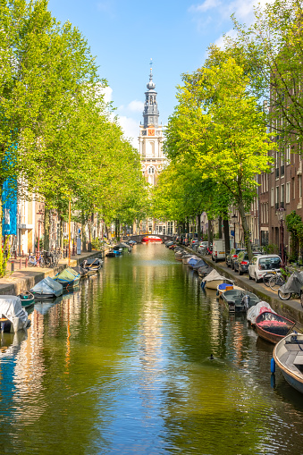 Netherlands. Sunny summer day on the Amsterdam canal. Parked cars on the embankment and many moored boats on the water. Cathedral building in the distance