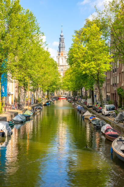 Amsterdam Canal with Boats on a Sunny Day Netherlands. Sunny summer day on the Amsterdam canal. Parked cars on the embankment and many moored boats on the water. Cathedral building in the distance canal house photos stock pictures, royalty-free photos & images