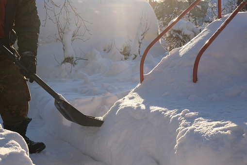 A man with a snow shovel removes snow in the yard in the light of the sun