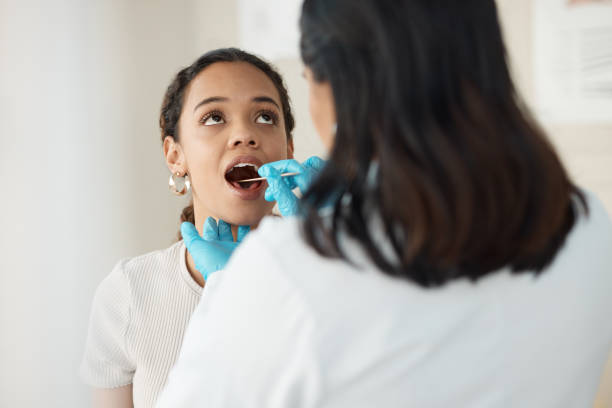 scatto di una giovane donna seduta in clinica mentre il suo medico le esamina la gola durante una consultazione - human mouth foto e immagini stock