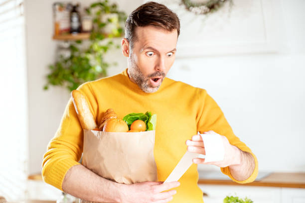 hombre sorprendido mirando el recibo de la tienda después de comprar, sosteniendo una bolsa de papel con alimentos saludables. chico en la cocina. - inflation fotografías e imágenes de stock