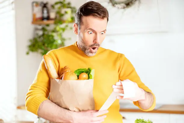 Surprised man looking at store receipt after shopping, holding a paper bag with healthy food. Guy in the kitchen. Real people expression. Inflation concept.