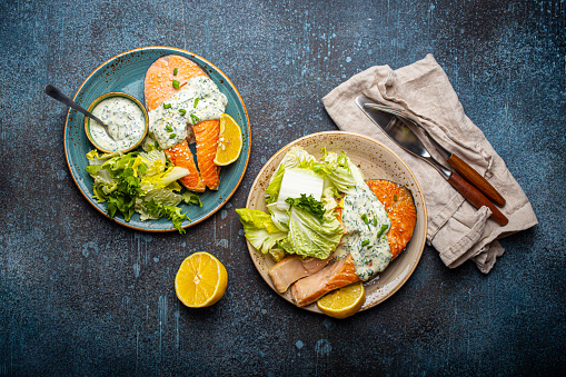 Healthy food meal grilled salmon steaks with dill sauce and salad leafs on two plates on rustic concrete stone background table flat lay from above, diet healthy nutrition dinner