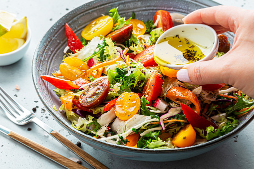 Fresh colorful spring vegetable salad with cherry tomatoes and sweet peppers in the blue bowl. Cookâs hand pouring olive oil with herbs (dressing). Healthy organic vegan lunch or snack close up.