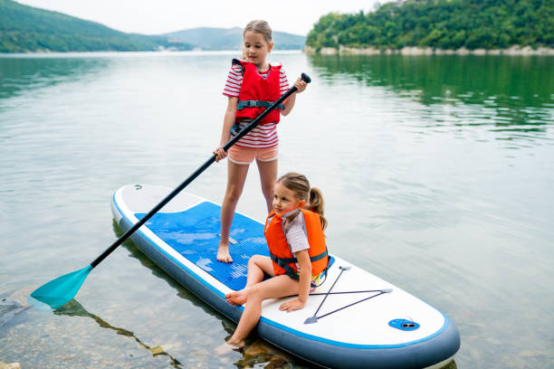 girls padding on stand up paddle boarding on lake district. children in swim life vest learning paddleboarding on sup board. family active leisure and local getaway with kids concept - paddle surfing stockfoto's en -beelden