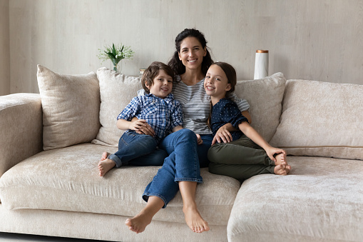 Family portrait smiling mother hugging little son and daughter, sitting on cozy couch at home, happy loving Hispanic young mom embracing adorable kids, looking at camera, posing for photo together