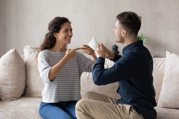 Smiling woman who is deaf and man using sign language Smiling young woman who is deaf and man using sign language, sitting on couch at home, excited beautiful girlfriend and boyfriend having fun, enjoying pleasant conversation, hard of hearing sign language stock pictures, royalty-free photos & images