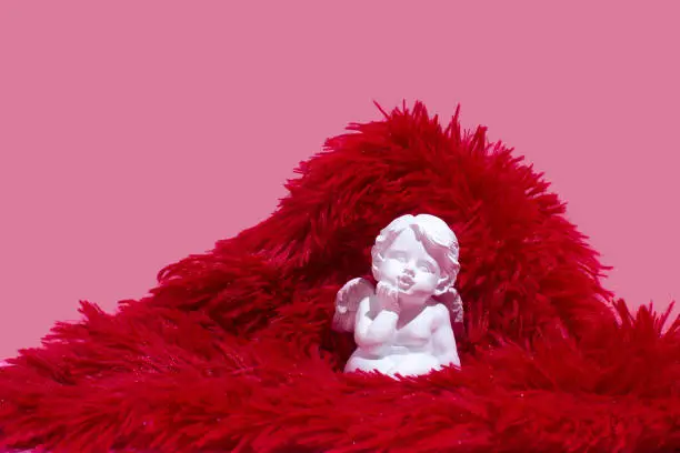 Photo of White angel statue with wings sitting on red soft shaggy fabric against pink background. St Valentine love minimal concept.