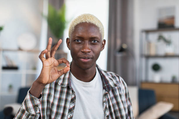 African man with blond hair gesturing sign OK with fingers Portrait of african man with blond hair and blue eyes gesturing sign OK with fingers while sitting at home and looking at camera. Concept of people and approvement. black men with blonde hair stock pictures, royalty-free photos & images