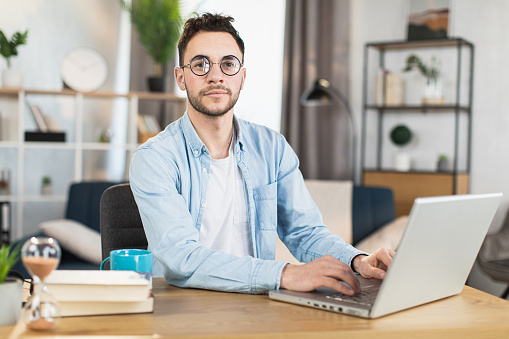 Handsome caucasian man in eyeglasses and casual wear looking at camera while sitting at desk with modern laptop. Concept of people, technology and freelance.