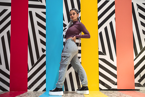 Full length view of a young woman with white braids standing and posing sideways with hand on waist while looking at camera against wall with colors and striped designs.