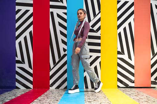 Full length view of a young woman with white braids standing and looking at camera against wall with colors and striped designs.