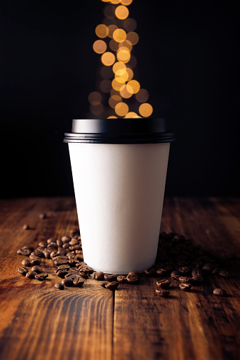 Paper cup of takeaway coffee with coffee beans on rustic wooden table and bokeh lights on background, Close up