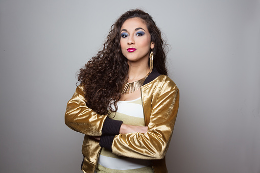 Studio portrait of a beautiful Hispanic woman with long curly hair and brown eyes, she has put on colorful make up and is wearing a golden jacket and huge  golden jewelry.