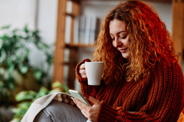 una linda chica de jengibre con cabello rizado está sentada en la silla de casa por la mañana, enviando mensajes de texto por teléfono y bebiendo su café. - young women sitting simple living eastern europe fotografías e imágenes de stock