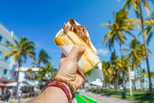 POV Point of view shot of a young travel male enjoying his vacations while eating a cuban sandwich in front of Ocean Drive, South Beach, Miami Beach, Miami, South Florida, United States of America.\n\nThe picture is in the famous and touristic Art Deco District. Shooting from a personal perspective in an exotic tropical beach travel holidays.\n\nTravel Concept.