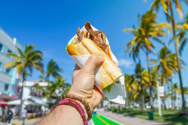 POV Point of view shot of a young travel male enjoying his vacations while eating a cuban sandwich in front of Ocean Drive, South Beach, Miami Beach, Miami, South Florida, United States of America.

The picture is in the famous and touristic Art Deco District. Shooting from a personal perspective in an exotic tropical beach travel holidays.

Travel Concept.