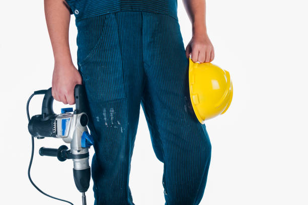 Young male construction worker with beard wearing red helmet and plaid shirt using power drill inside wooden house under construction indoors with natural light and visible wooden beams
