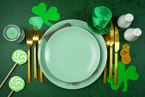 Happy St Patrick's Day plates table setting, styled with leprechaun hat, shamrocks, and chocolate gold coins, on a textured green background. Mockup. Top view flat lay. Copy space.