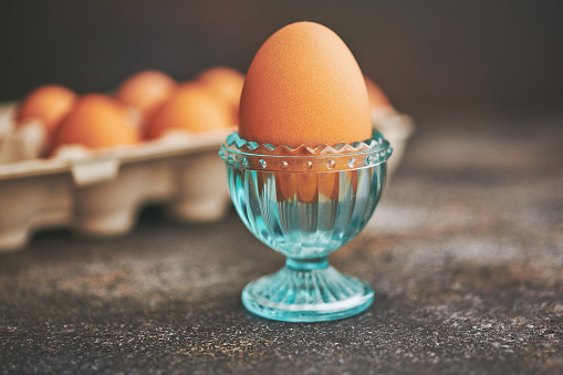 Fresh organic boiled egg in a crystal eggcup with brown eggs in a cardboard egg carton in the background