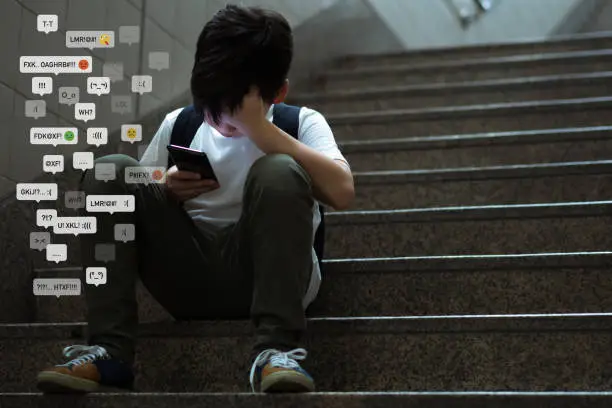 Photo of Asian teenage boy sitting at stair, covering his face with hands, face down, holding smartphone in low light.