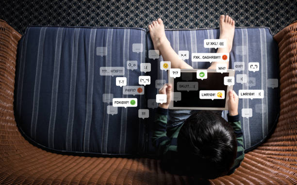 Top view of little Asian boy child using computer tablet alone on sofa in dark room with cyberbullying emoticon. Table top view of little Asian boy child using computer tablet alone on sofa in dark room with emoticon. Cyberbullying, social media addiction, and hate speech concept. cyberbullying stock pictures, royalty-free photos & images