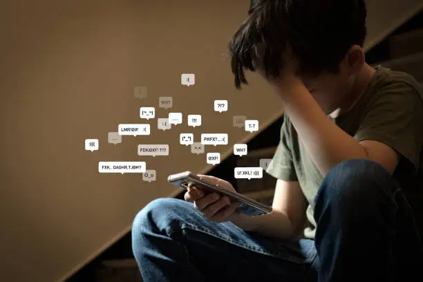 Asian teenage boy sitting at stair, covering his face with hands, face down, holding smartphone in low light feeling frustrated, lonely, stress and depressed. Cyber bullying concept.