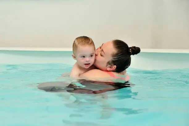 A mother and her child with Down syndrome swim and play in a children's pool with blue water. A woman hugs and kisses her disabled baby tightly. Portrait. Horizontal orientation.