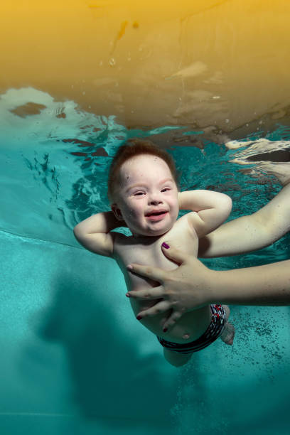 un petit garçon heureux atteint du syndrome de down dans les bras d’un parent sous l’eau dans une piscine d’eau turquoise. l’enfant regarde dans la caméra avec ses mains derrière la tête et sourit. orientation verticale. - portrait babies and children people nature photos et images de collection