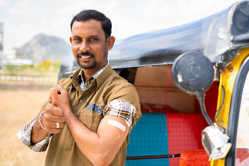 Auto driver showing thumbs up with coronavirus or covid-19 vaccinated bandage on shoulder by looking at camera - concept of vaccination recommendation, responsibility and prevention from infection.