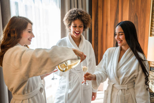 Excited Mixed Race Friends Celebrating With Champagne And Having Fun In A Hotel Room Diverse young women laughing, dancing and relaxing with a glass of sparkling wine. bathrobe photos stock pictures, royalty-free photos & images