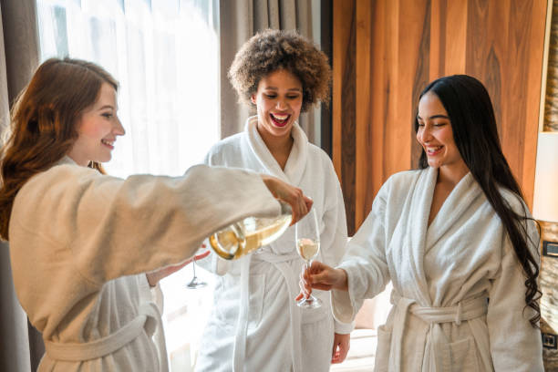 Excited Mixed Race Friends Celebrating With Champagne And Having Fun In A Hotel Room Diverse young women laughing, dancing and relaxing with a glass of sparkling wine. spa lady stock pictures, royalty-free photos & images