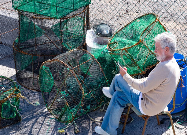 istock White-haired elderly fisherman on the dock of the harbor relaxes after work using his mobile phone, sitting near the fishing nets to prepare 1369702541