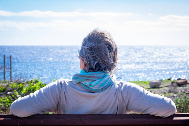 vista traseira de uma mulher madura de cabelos grisalhos relaxando na frente do mar, sentada em um banco em um dia de vento. idosa desfrutando de aposentadoria olhando o horizonte sobre a água - wind scarf women people - fotografias e filmes do acervo