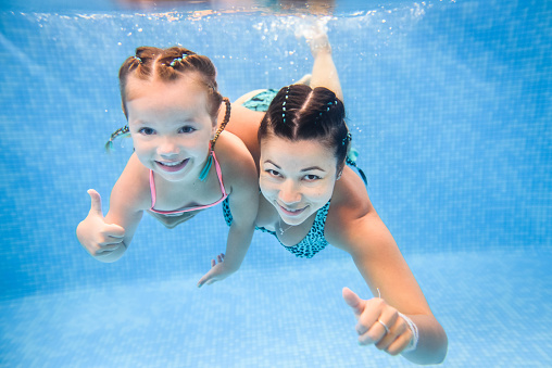 Mom and daughter are immersed in water, swimming under water in a paddling pool. Diving kid. Learning child to swim. Young mother or swimming instructor and happy little girl.