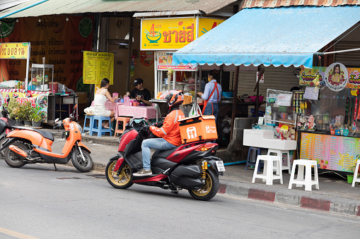 Thai delivery person on motorcycle is leaving street restaurant on street Chikchai 4 in Bangkok Ladprao. In background people are sitting and eating at restaurant