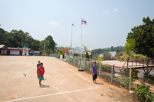 View onto school courtyard and sports playground in town Pang Chum in north Thailand in Amphoe Samsoeng in Chiang Mai province. A few thai teengares are playing barefeet and walking in sparetime.  At right center  are flag poles and thai letters.