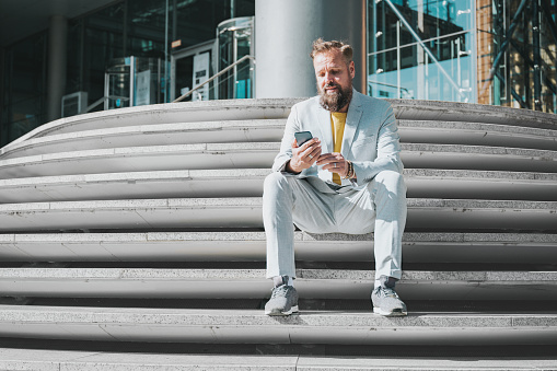 elegant bearded business man sitting outdoors on stairs in front of office in Berlin and tapping at mobile