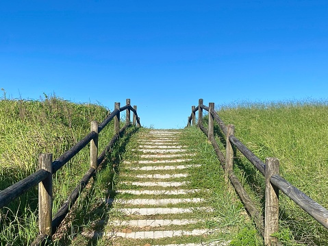 Horizontal vanishing point of walking up sandy green grass wood fence post lined stairs leading to beach coastline with sand dunes under a clear blue sky day Australia